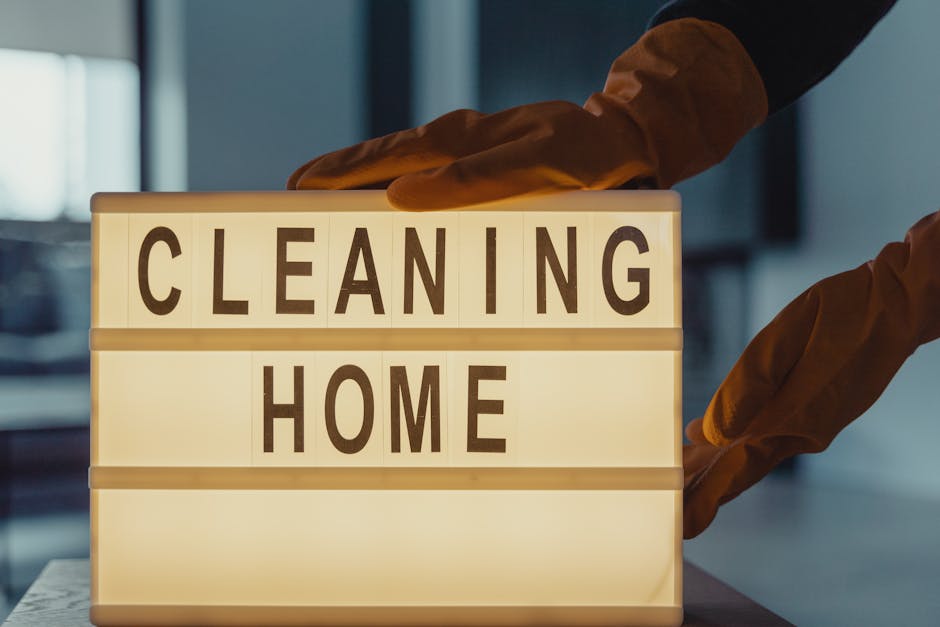 A person wearing orange cleaning gloves holds a lit-up message board displaying the words 'CLEANING HOME' in bold black letters. The background shows a modern, well-lit interior with a blurred view of kitchen or living space. The message board emphasizes domestic cleaning services, aligning with surface cleaning and sanitisation practices promoted by Carpet Cleaners Ilford, as detailed on their website page about avoiding hidden cleaning charges in Ilford.