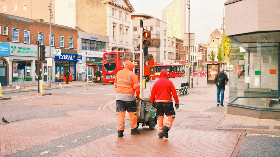 Two street cleaning workers wearing orange and red high visibility jackets and trousers are pushing a cart with cleaning tools along a paved sidewalk in Ilford. The scene includes a crosswalk with tactile paving, nearby benches, and a bus stop. In the background, red double-decker buses are visible along with commercial buildings including a store labeled 'Coral' and a pharmacy. The area appears clean and well-maintained, with bright natural lighting illuminating the urban setting. The image is associated with Carpet Cleaners Ilford, emphasizing surface and deep cleaning services for maintaining hygiene and presentation in residential and commercial spaces.