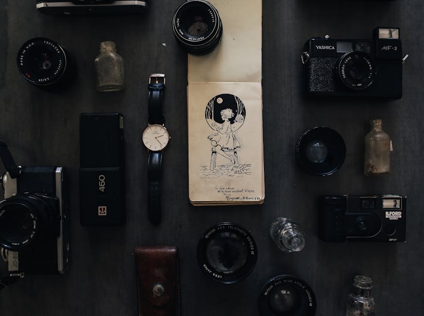 An overhead view of a dark wooden surface featuring various vintage and modern camera equipment, including a Yashica camera, a black film camera, and multiple camera lenses. There are also small glass bottles, a black external hard drive, a wristwatch, and a sketchpad displaying an illustration of a girl sitting on a dock at night. The surface is clean and well-organized, with soft lighting highlighting the condition and details of each item, indicative of a photography enthusiast's collection. The arrangement demonstrates attention to surface cleanliness and object care, aligning with professional domestic or surface cleaning practices offered by Carpet Cleaners Ilford, especially for delicate or valuable items.