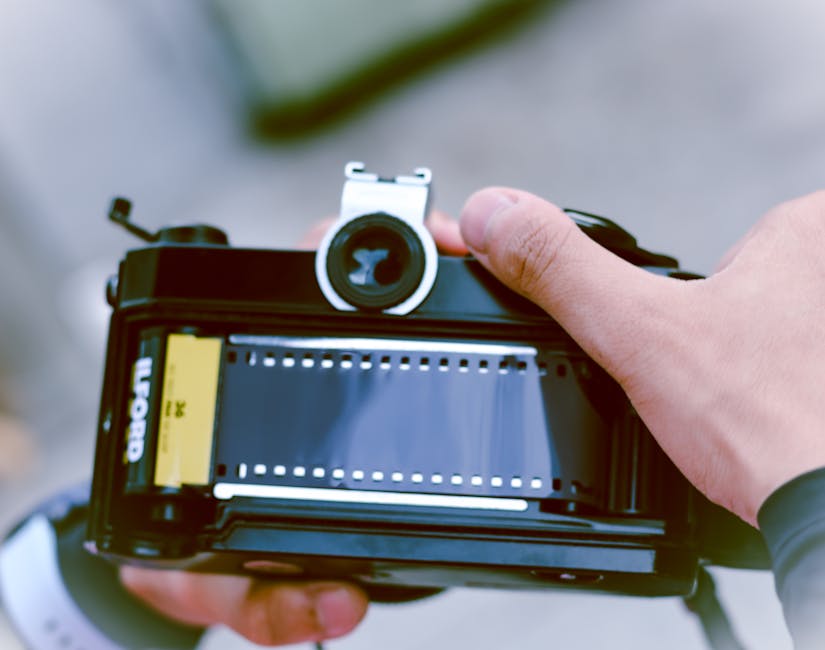 A close-up of a person holding a professional film camera with a black body and silver lens, positioned horizontally outdoors. The person's hand is gripping the camera's body, with visible fingers and a wristwatch. The camera features a yellow label on the side and appears to be set for filming. In the background, there is a blurred outdoor scenery with soft lighting, emphasizing the focus on the camera used in professional filming or video production, reflecting attention to detail and equipment quality consistent with high-standard media work. This image relates to content about surface cleaning and maintenance, highlighting the importance of meticulous attention to detail in the cleaning process, similar to precise camera work, and referencing Carpet Cleaners Ilford for domestic cleaning services.