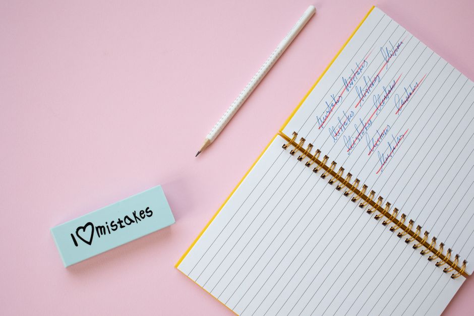 A pink surface with an open spiral-bound notebook displaying handwritten notes in blue and red ink, a white pencil with a pink eraser placed diagonally above the notebook, and a small light blue card with the handwritten message 'I ❤️ mistakes' in black ink and a heart drawing. The scene emphasizes themes of reflection and accuracy, relevant to proper cleaning practices and avoiding common booking errors for Ilford cleaning appointments, as highlighted on the Carpet Cleaners Ilford website.