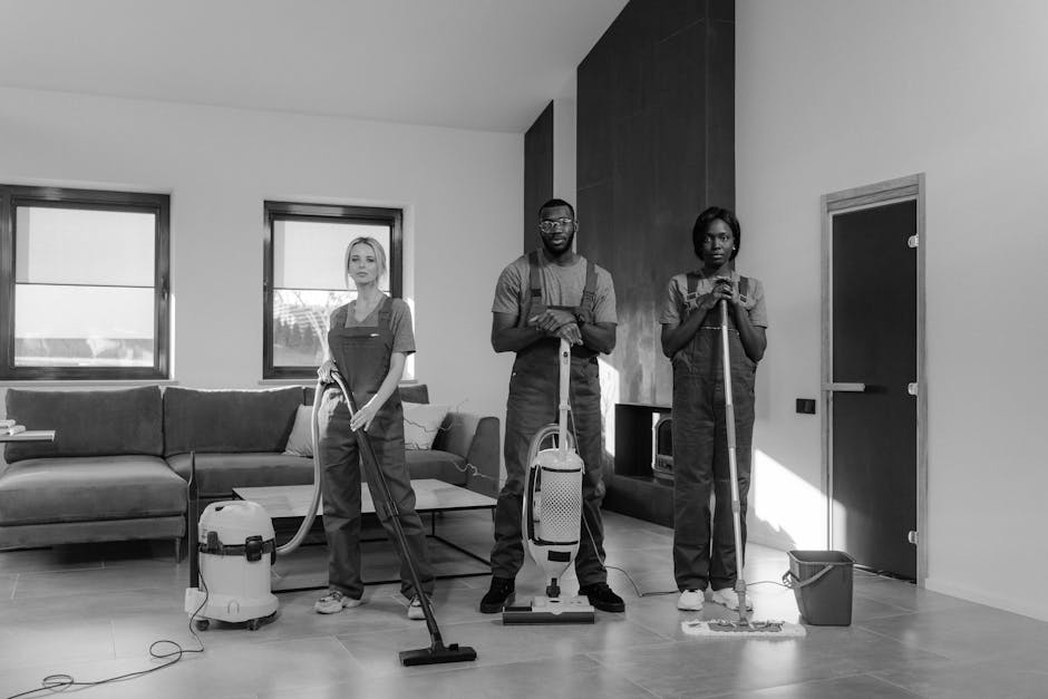 A professional cleaning team comprising three individuals is performing surface cleaning in a modern living room. The room features a light-colored tiled floor, a dark fabric sofa, and a coffee table. Two large windows on the left allow natural light to illuminate the space, highlighting the clean and dust-free condition of the polished floor. The team members are dressed in uniforms with aprons—one holding a vacuum cleaner, another with a carpet scrubber, and the third with a mop and bucket—demonstrating various deep cleaning and sanitisation tools used in residential or commercial cleaning. Visible fixtures include a wall-mounted device, possibly a thermostat or light switch, and an enclosed cabinet on the right. Overall, the scene emphasizes meticulous cleaning processes, aligning with the themes of gentle surface care and hygiene maintained by Carpet Cleaners Ilford in their professional domestic cleaning services, as highlighted in the page about common booking mistakes for Ilford cleaning appointments.