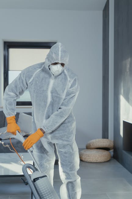 A professional wearing a white protective suit, face mask, and orange gloves using a portable vacuum cleaner for deep cleaning or sanitisation in a modern indoor setting, likely a residential or commercial space. The room features grey tiled flooring, large windows allowing natural light, and a minimalistic decor including small round cushions on the floor near a dark wall. The scene emphasizes surface cleaning and hygiene maintenance, aligned with services offered by Carpet Cleaners Ilford, with a focus on thorough sanitisation and deep cleaning to ensure a hygienic environment.