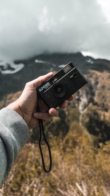 A person holding a black point-and-shoot camera with a wrist strap, aimed at a foggy landscape with distant mountains and sparse vegetation, under overcast skies. The person's hand and part of their grey sleeve are visible, while the background features a mist-covered terrain with rocky hills and cloudy weather, creating a moody, atmospheric scene.