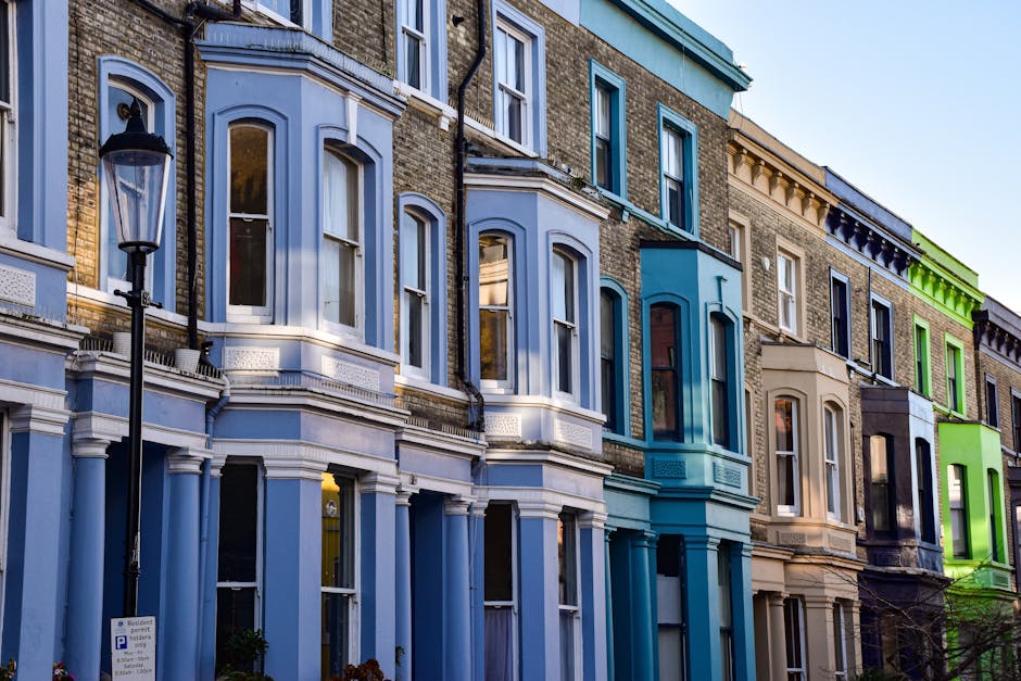A row of colorful Victorian-style terraced houses in Ilford, London, featuring bay windows, brick facades, and decorative cornices. The buildings are painted in pastel shades of blue, beige, cream, and vibrant green, with large, clear glass windows reflecting the daylight. The street scene includes a classic black street lamp, and the houses appear well-maintained with clean exteriors. The bright sky and daylight highlight the architectural details and fresh appearance of the facades, illustrating a typical residential street in the Ilford area, enhanced by the presence of a sign indicating parking regulations. This image exemplifies well-kept urban housing, ideal for illustrating neighbourhood cleanliness, property maintenance, or domestic cleaning references in the context of Gants Hill end of tenancy cleaning guide for Ilford landlords, provided by Carpet Cleaners Ilford.