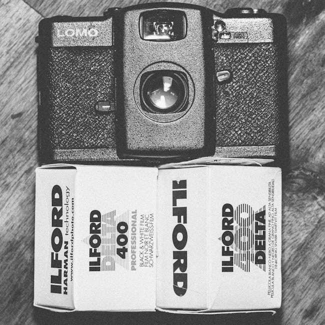 A black and white photograph showing a vintage film camera positioned above a box of Ilford HP5 Plus 400 professional black and white film. The camera is resting on a wooden surface, with the lens facing downward. The camera body features textured grips on the sides and a prominent lens in the center. The film box, placed directly below the camera, displays the Ilford logo and product information in bold lettering. The scene is evenly lit, emphasizing the textures of the camera and the grain of the wooden surface, reflecting a focus on classic photographic equipment which could be related to detailed cleaning and maintenance of sensitive surfaces. This setup illustrates a clean, organized workspace suitable for professional cleaning or camera care, consistent with the services provided by Carpet Cleaners Ilford in the context of surface cleaning and maintenance.