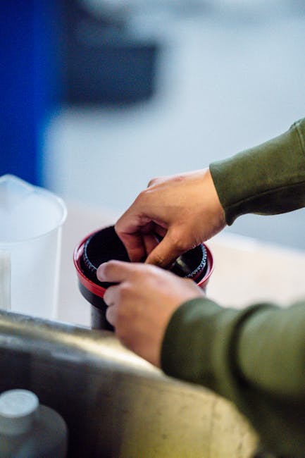 A person wearing a green long-sleeved shirt is cleaning a black and red cylindrical container, possibly a vacuum filter or cleaning tool, over a light-colored kitchen or laundry sink. The surface around the sink is smooth and appears to be made of stainless steel or a similar material, with a white plastic container nearby. Bright natural or artificial lighting illuminates the countertop, emphasizing a clean and tidy environment. This image illustrates detailed surface cleaning processes, which are part of domestic cleaning practices supported by Carpet Cleaners Ilford, a professional cleaning service near Ilford Station IG1. The focus on the hands handling a cleaning accessory highlights thorough maintenance and hygiene efforts.