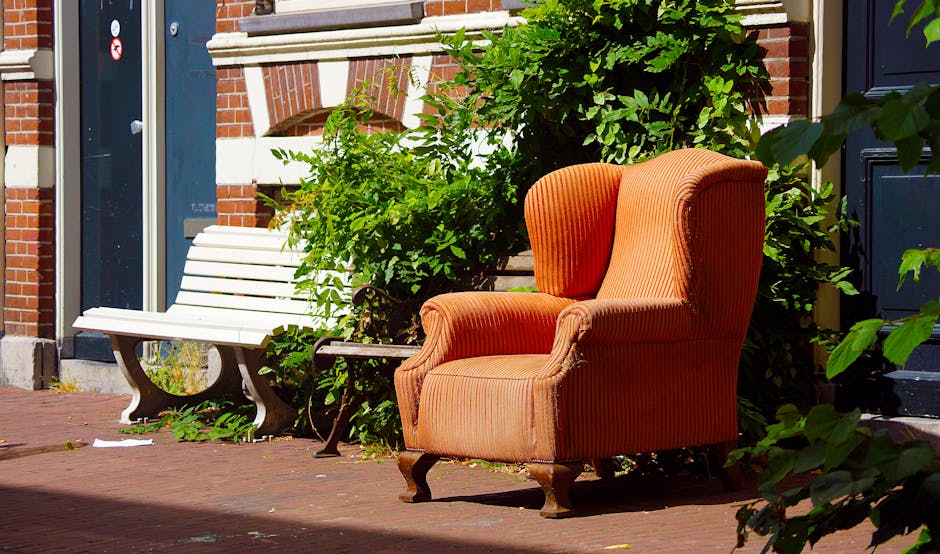A red leather armchair is shown outdoors, leaned against a rough stone wall on a paved walkway. The chair appears to be discarded, with visible signs of wear and dirt on its surface. Surrounding the c