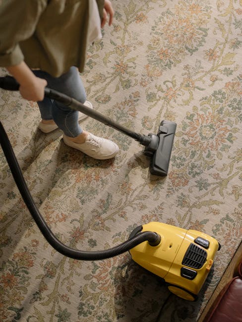 A person using a yellow vacuum cleaner with a black hose attachment on a traditional patterned area rug in a domestic setting. The rug features a floral design with beige, green, and reddish tones. The person, wearing beige shoes, blue jeans, and a light-colored jacket, is standing near a piece of wooden furniture. Bright lighting illuminates the room, emphasizing the clean and well-maintained appearance of the surface. Carpet Cleaners Ilford is visible, indicating a professional cleaning service specializing in deep surface cleaning and sanitisation for residential spaces.